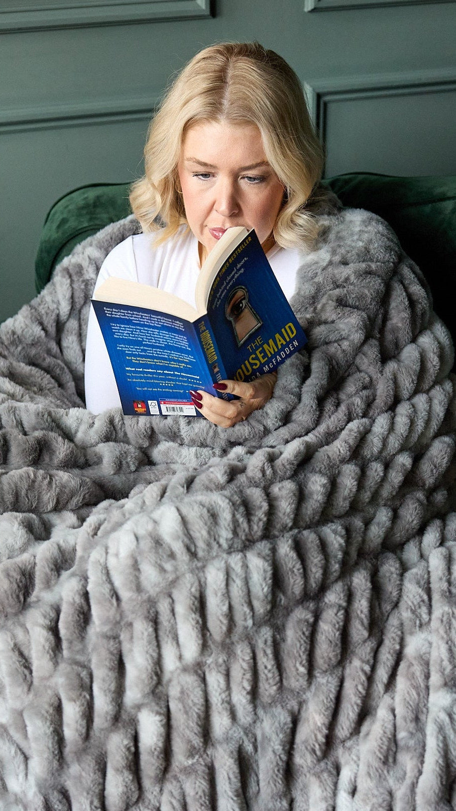 Woman reading a book wrapped in a gray blanket on a green couch.
