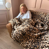 A woman smiling while sitting on a tan leather sofa and wrapped in a plush leopard-print throw blanket in a softly lit room with blush pink walls.