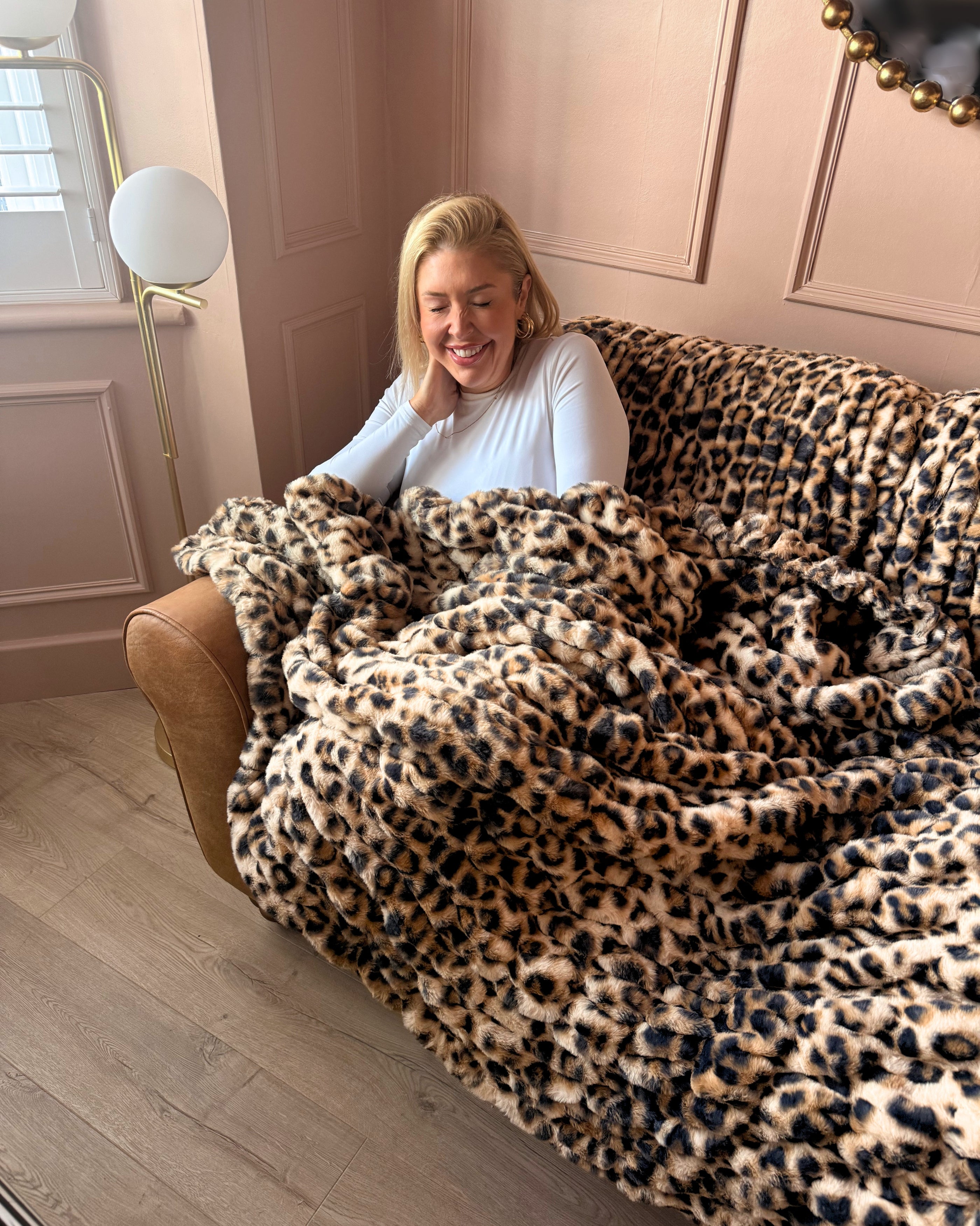 A woman smiling while sitting on a tan leather sofa and wrapped in a plush leopard-print throw blanket in a softly lit room with blush pink walls.