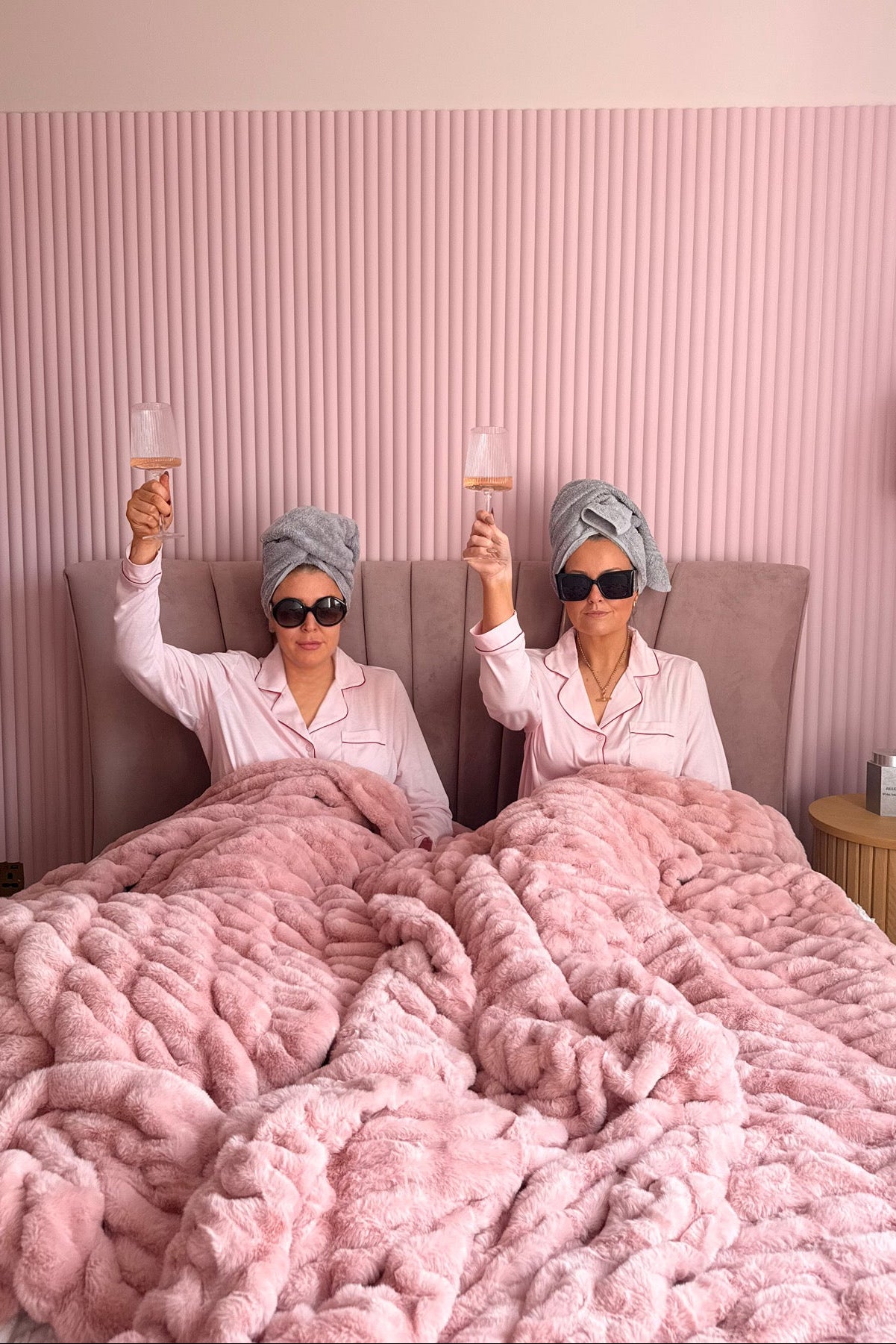 Two women in bed with pink blankets, wearing turmeric and sunglasses, holding drinking glasses.