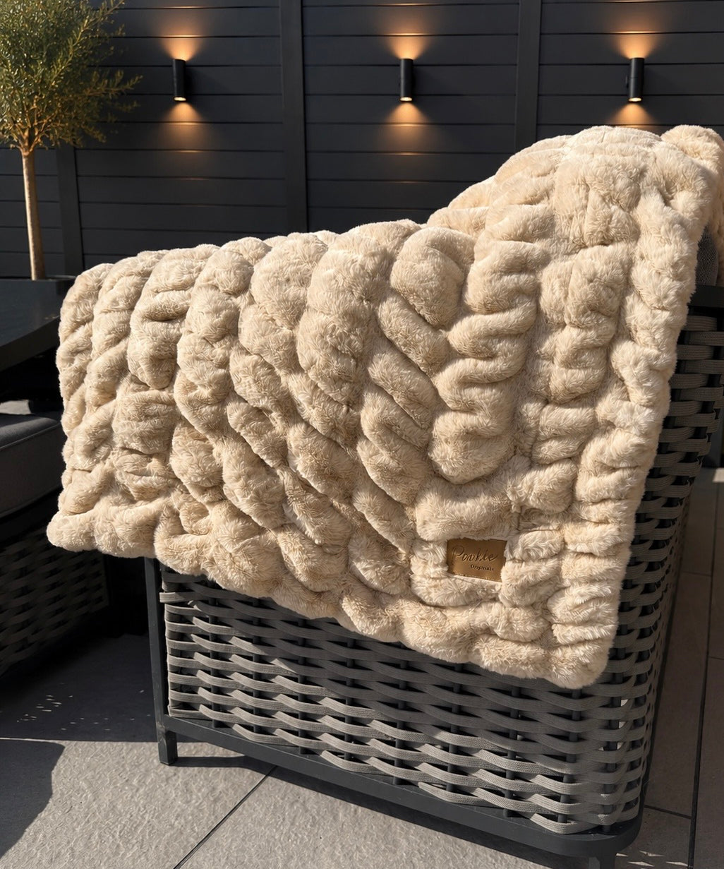 Beige textured blanket draped over a wicker chair outdoors with a dark fence and plants in the background.