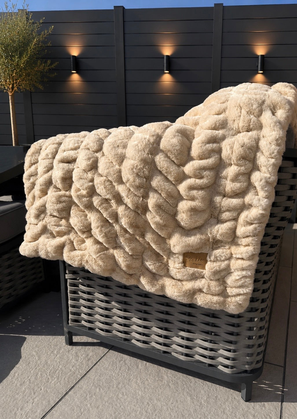 Beige textured blanket draped over a wicker chair outdoors with a dark fence and plants in the background.