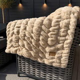 Beige textured blanket draped over a wicker chair outdoors with a dark fence and plants in the background.